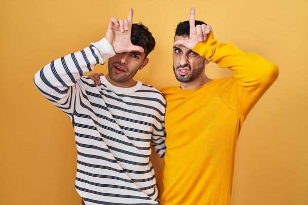 Νέοι ισπανόφωνοι γκέι ζευγάρι που στέκονται πάνω από κίτρινο φόντο κοροϊδεύοντας ανθρώπους με δάχτυλα στο μέτωπο κάνοντας αποτυχημένες χειρονομίες χλευάζοντας και προσβάλλοντας.  - Φωτογραφία, εικόνα