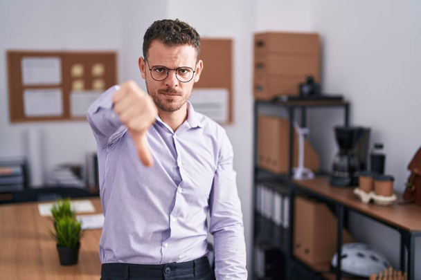 Νεαρός Ισπανός στο γραφείο δείχνει δυστυχισμένος και θυμωμένος δείχνοντας απόρριψη και αρνητικός με τους αντίχειρες κάτω. κακή έκφραση.  - Φωτογραφία, εικόνα