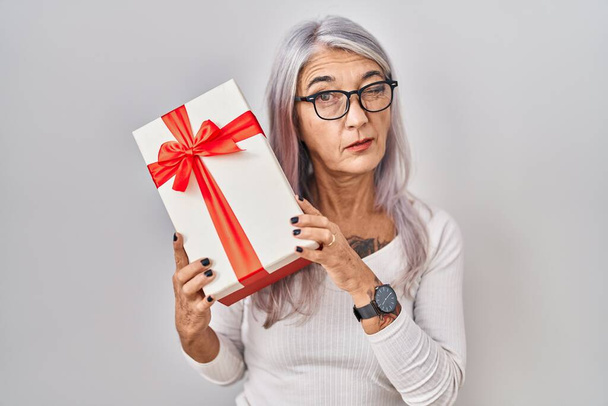 Žena středního věku s šedými vlasy drží dárek mrkání na kameru se sexy výraz, veselý a šťastný obličej.  - Fotografie, Obrázek