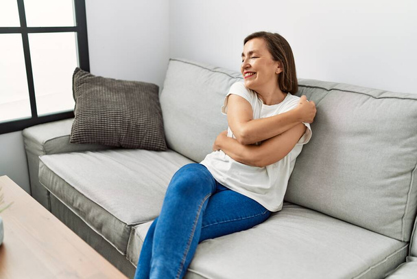 Middle age hispanic woman smiling sitting on the sofa hugging herself at home - Fotó, kép