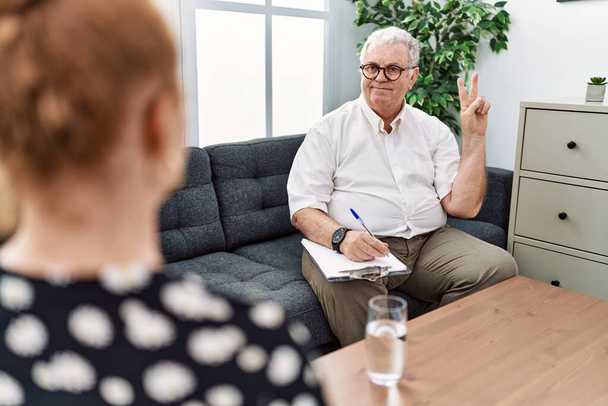 Senior psychologist man at consultation office smiling with happy face winking at the camera doing victory sign with fingers. number two.  - Фото, изображение