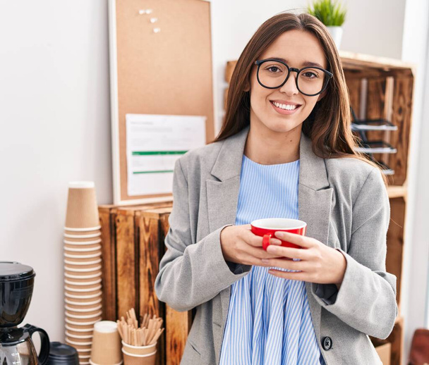 Νεαρή όμορφη ισπανόφωνη επιχειρηματίας χαμογελά αυτοπεποίθηση πίνοντας καφέ στο γραφείο - Φωτογραφία, εικόνα