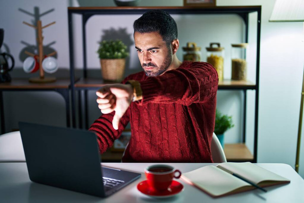 Fiatal, szakállas spanyol férfi számítógép laptoppal éjjel otthon boldogtalannak és dühösnek tűnik, elutasítást mutat, és negatív hüvelykujjal lefelé gesztussal. rossz kifejezés.  - Fotó, kép