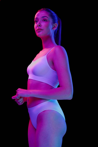 Portrait of young beautiful woman posing in white underwear isolated over black background in neon light. Looking away. Concept of beauty, body and skin care, health, plastic surgery, cosmetics - Фото, изображение