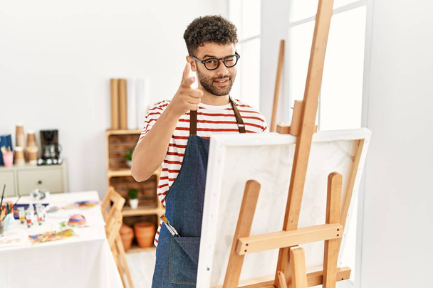 Арабський молодий чоловік у художній студії вказує пальцями на камеру з щасливим і кумедним обличчям. добра енергія й трубопроводи.  - Фото, зображення