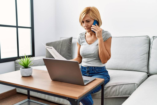 Middle age blonde woman talking on the smartphone using laptop at home - Foto, Imagem