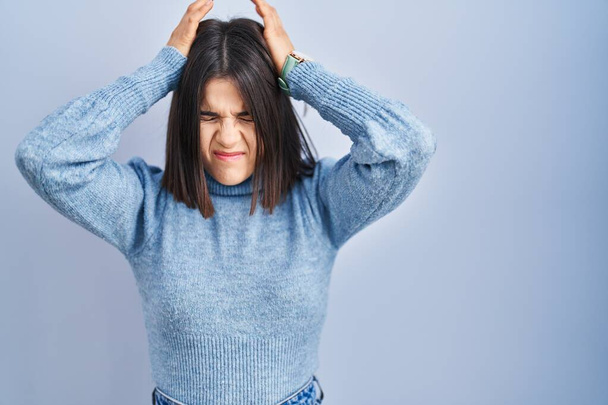 Nuori latino nainen seisoo sininen tausta kärsii päänsärky epätoivoinen ja korosti, koska kipu ja migreeni. kädet pään päällä.  - Valokuva, kuva