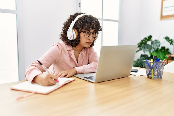 Νεαρή επιχειρηματίας της Μέσης Ανατολής που χρησιμοποιεί ακουστικά και laptop γράφοντας στο ημερολόγιο εργασίας στο γραφείο. - Φωτογραφία, εικόνα