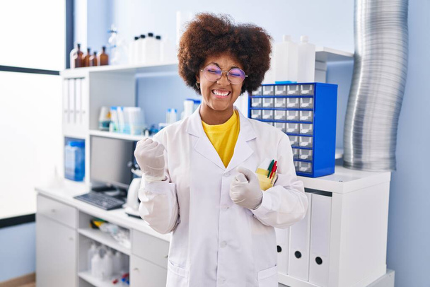 Νεαρή Αφροαμερικανή που εργάζεται σε εργαστήριο επιστημόνων γιορτάζει έκπληκτος και έκπληκτος για την επιτυχία με τα χέρια ψηλά και τα μάτια κλειστά  - Φωτογραφία, εικόνα
