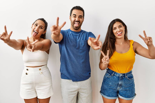 Een groep jonge Latijns-Amerikaanse mensen die over een geïsoleerde achtergrond staan te glimlachen met hun tong en vingers laten zien van beide handen die een overwinningsteken doen. nummer twee.  - Foto, afbeelding