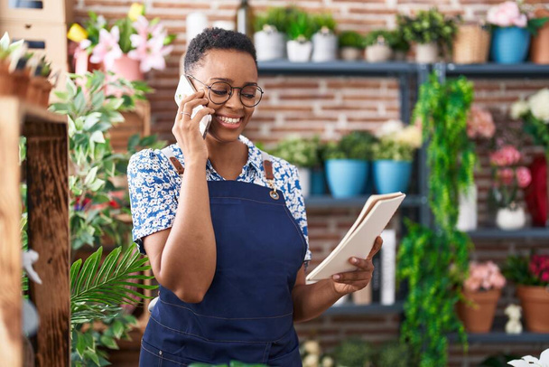 African american woman florist talking on smartphone reading notebook at florist - Photo, Image