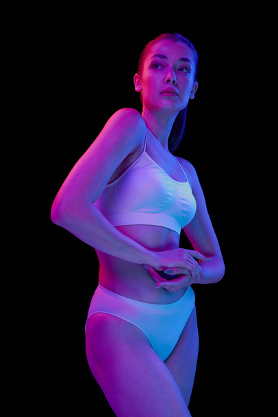 Portrait of young beautiful woman posing in white underwear isolated over black background in neon light. Concept of beauty, body and skin care, health, plastic surgery, cosmetics - Fotó, kép