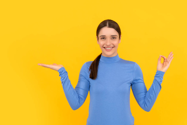 advertisement. cheerful woman presenting product on copy space. beauty and fashion. female fashion model. young girl show ok. portrait of lady on yellow background. express positive emotions. - Photo, Image