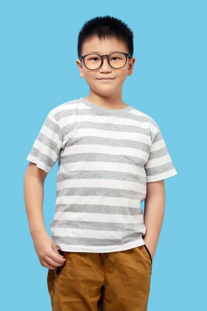 Enfant intelligent debout avec le sourire, portant des lunettes et un t-shirt regardant la caméra sur fond bleu - Photo, image