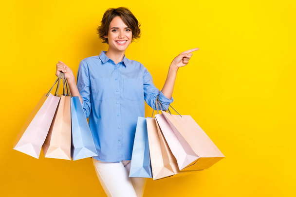 Portrait of lovely girl with bob hairstyle wear blue blouse hold shopping bags directing empty space isolated on yellow color background. - Foto, immagini