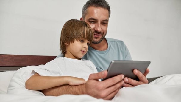 Father and little son watching digital tablet in bed at home. Young caucasian family relationship. Man hugging boy. Fatherhood and parenting. Domestic leisure, hobby and entertainment - Фото, изображение