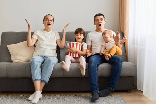 Excited woman and man wearing casual white t-shirts sitting on sofa with their children and watching something amazing on tv, looking with big eyes, saying wow. - Фото, изображение