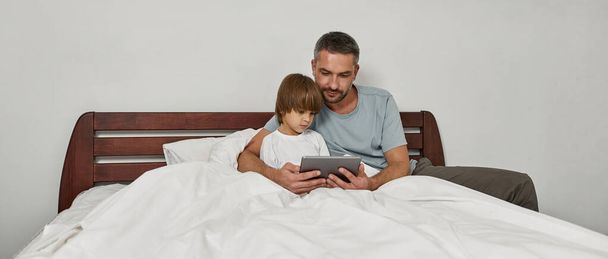 Focused father and little son watching digital tablet in bed at home. Young european family relationship and spending time together. Fatherhood and parenting. Domestic leisure, hobby and entertainment - 写真・画像