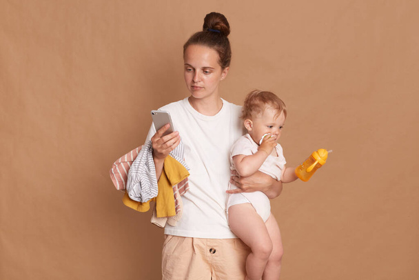Indoor shot of serious young adult woman with bun hairstyle wearing white t shirt standing with her toddler daughter and using mobile phone with concentrated expression, isolated over brown background - Foto, Bild