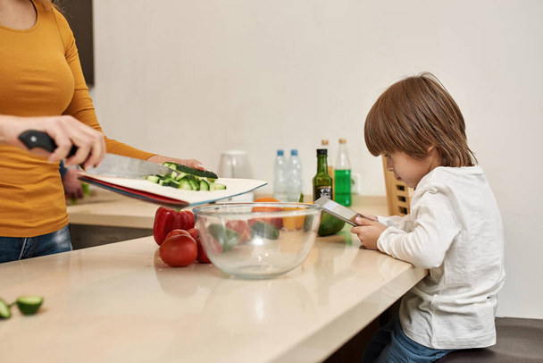 European little boy using digital tablet while his cropped mother pouring cutting cucumber in bowl for salad at home kitchen. Parenting. Family relationship and spend time together. Domestic lifestyle - Φωτογραφία, εικόνα