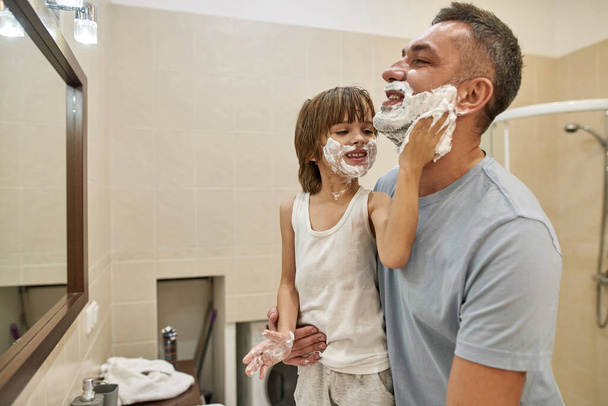 Little boy smearing shaving foam on father beard at home bathroom. Side view of smiling caucasian child and man joking and spending time together. Fatherhood and parenting. Face skin care and hygiene - Fotoğraf, Görsel
