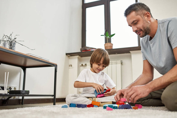 Pa en zoontje spelen thuis met bouwstenen op tapijt. Blanke familierelatie en samen tijd doorbrengen. Vaderschap en ouderschap. Huishoudelijke vrijetijdsbesteding, hobby en entertainment - Foto, afbeelding