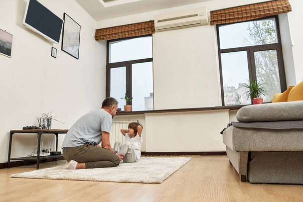 Father teaching little son doing press ups on carpet at home. Man and boy look at each other. Young european family relationship and spending time together. Fatherhood and parenting. Healthy lifestyle - Фото, зображення