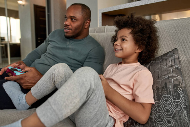 Black little boy and father playing video game with joysticks on sofa at home. Smiling child and concentrated man enjoying time together. Fatherhood and parenting. Domestic leisure and entertainment - 写真・画像