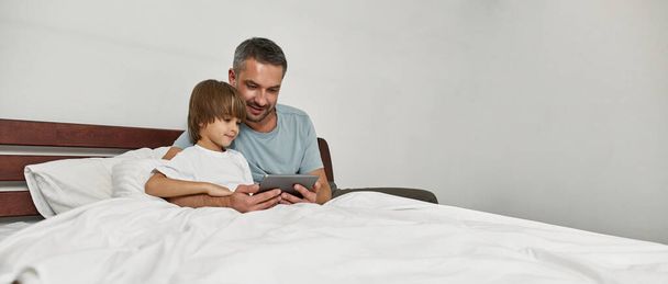 Concentrated father and little son watching digital tablet in bed at home. Caucasian family relationship and spending time together. Fatherhood and parenting. Domestic leisure, hobby and entertainment - Foto, afbeelding