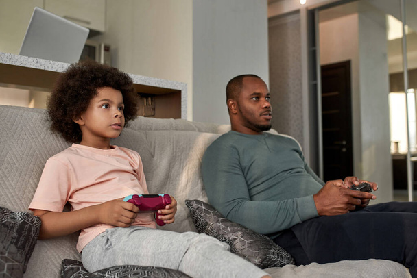 Black little boy and father playing video game with joysticks on sofa at home. Focused child and man spending time together. Fatherhood and parenting. Domestic leisure and entertainment - 写真・画像