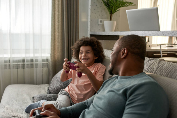 Black adult father look at little son during they play video game with joysticks on sofa at home. Smiling kid and man spend time together. Fatherhood and parenting. Domestic leisure and entertainment - Foto, imagen