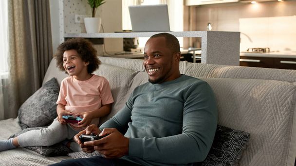 Black little boy and father playing video game with joysticks on sofa at home. Smiling and excited child and man enjoying time together. Fatherhood and parenting. Domestic leisure and entertainment - Fotó, kép