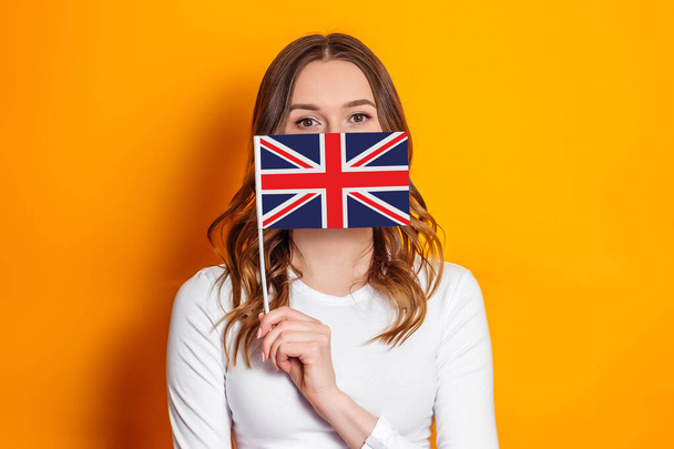Bildung im Ausland. Studentin bedeckt ihr Gesicht mit britischer Flagge auf orangefarbenem Hintergrund - Foto, Bild