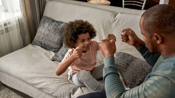 Little boy and adult father bumping fists of each other on sofa at home. Black man and smiling child playing and spending time together. Fatherhood and parenting. Domestic leisure and entertainment - Фото, зображення