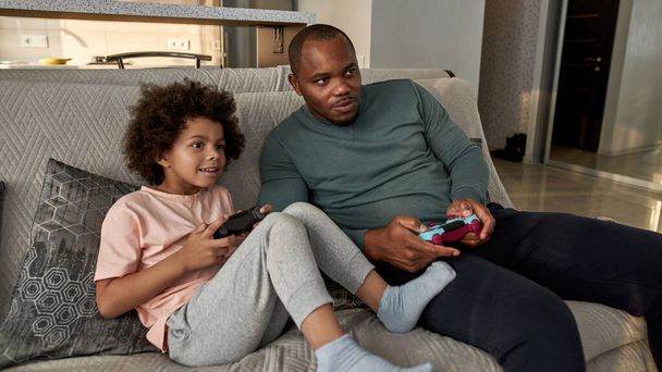 Black little boy and father playing video game with joysticks on sofa at spacious home. Child and man spending time together. Fatherhood and parenting. Domestic leisure and entertainment - Photo, Image
