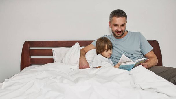 Father and little son reading book in bed at home. Young caucasian family relationship and spending time together. Fatherhood and parenting. Domestic leisure, hobby and entertainment - Zdjęcie, obraz