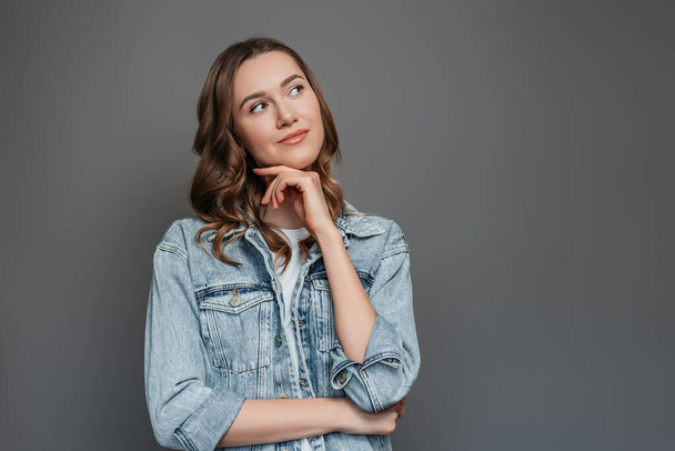 Female student in a denim jacket is thoughtful and stands isolated on a gray background in the studio, copy space - Foto, Bild