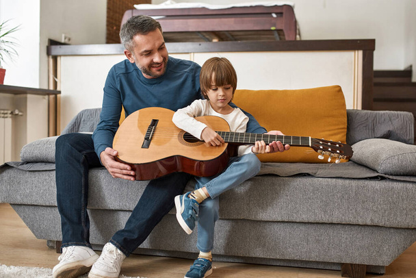 Smiling father teaching little son playing acoustic guitar on sofa at home. European family relationship and spending time together. Fatherhood and parenting. Domestic leisure, hobby and entertainment - Photo, image