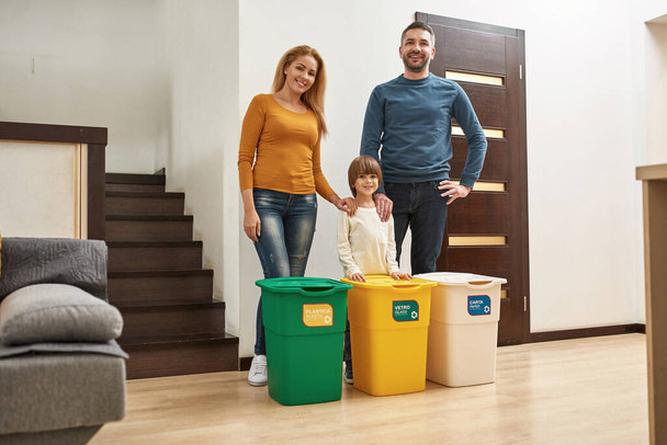 Young happy caucasian family standing near three dustbins for different trash at home. Ecological garbage recycle. Parenting. Father, mother and little son looking at camera. Modern domestic lifestyle - Фото, изображение