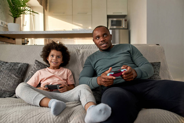 Curly black little boy and father playing video game with joysticks on sofa at home. Smiling child and focused man spending time together. Fatherhood and parenting. Domestic leisure and entertainment - Fotografie, Obrázek