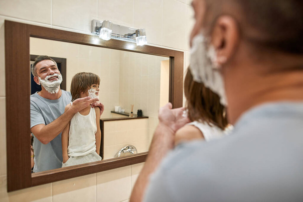 Mirror reflection of father smearing shaving foam on little son face at home bathroom. Caucasian child and excited man spending time together. Fatherhood and parenting. Face skin care and hygiene - Fotoğraf, Görsel