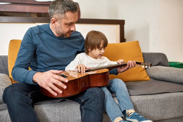 Father teaching little son playing acoustic guitar on sofa at home. Young caucasian family relationship and spending time together. Fatherhood and parenting. Domestic leisure, hobby and entertainment - Fotoğraf, Görsel