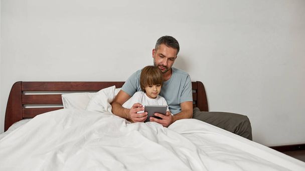 Father and little son watching digital tablet in bed at home. Young caucasian family relationship and spending time together. Fatherhood and parenting. Domestic leisure, hobby and entertainment - Photo, image