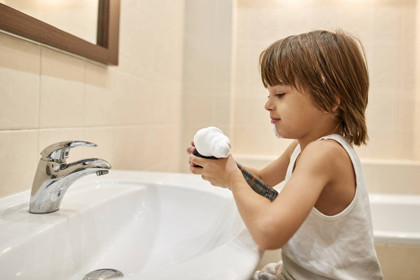 Little boy squeezing shaving foam from bottle over washing basin at home bathroom. Caucasian independent child teaching shaving. Modern domestic lifestyle. Face skin care and hygiene - Photo, Image