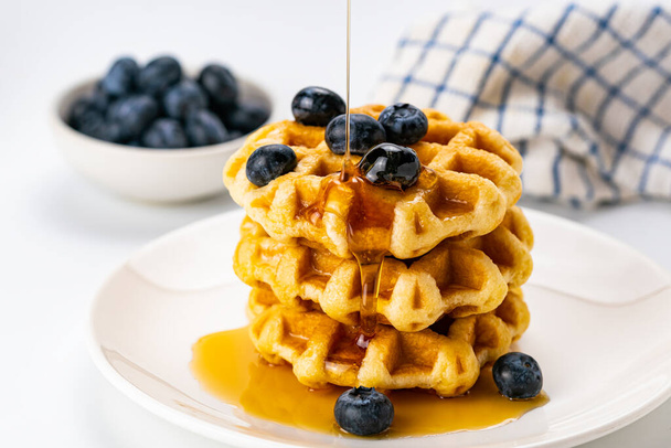 Closeup view of pouring honey on stack of fresh delicious homemade  waffles  topping with ripe organic blueberry fruit in white ceramic dish with blueberry in white ceramic cup and dish cloth on white background. - Photo, image