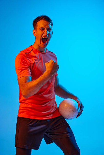 Happy emotions. Young male volleyball player with ball shouting isolated on blue studio background in neon light. Concept of summer sports, gym, team sport, challenges. - Fotografie, Obrázek