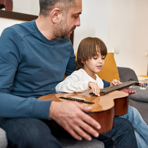 Father teaching little son playing notes on chords on acoustic guitar on sofa at home. Young caucasian family relationship. Fatherhood and parenting. Domestic leisure, hobby and entertainment - Фото, изображение