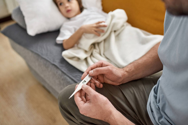 Selective focus of thermometer in hands of cropped father sitting on sofa near blurred lying sick little son with heat at home. Fatherhood and parenting. Health care. Family relationship - Foto, imagen