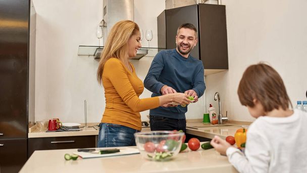 Man putting salad leaves on plate in hands of woman while their little boy using digital tablet at home kitchen. Parenting. Young caucasian family relationship. Modern domestic lifestyle - Photo, image
