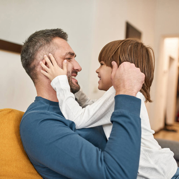 Father and little son looking and touching faces of each other on sofa at home. Young caucasian family relationship. Man and boy enjoying time and having fun together. Fatherhood and parenting - 写真・画像